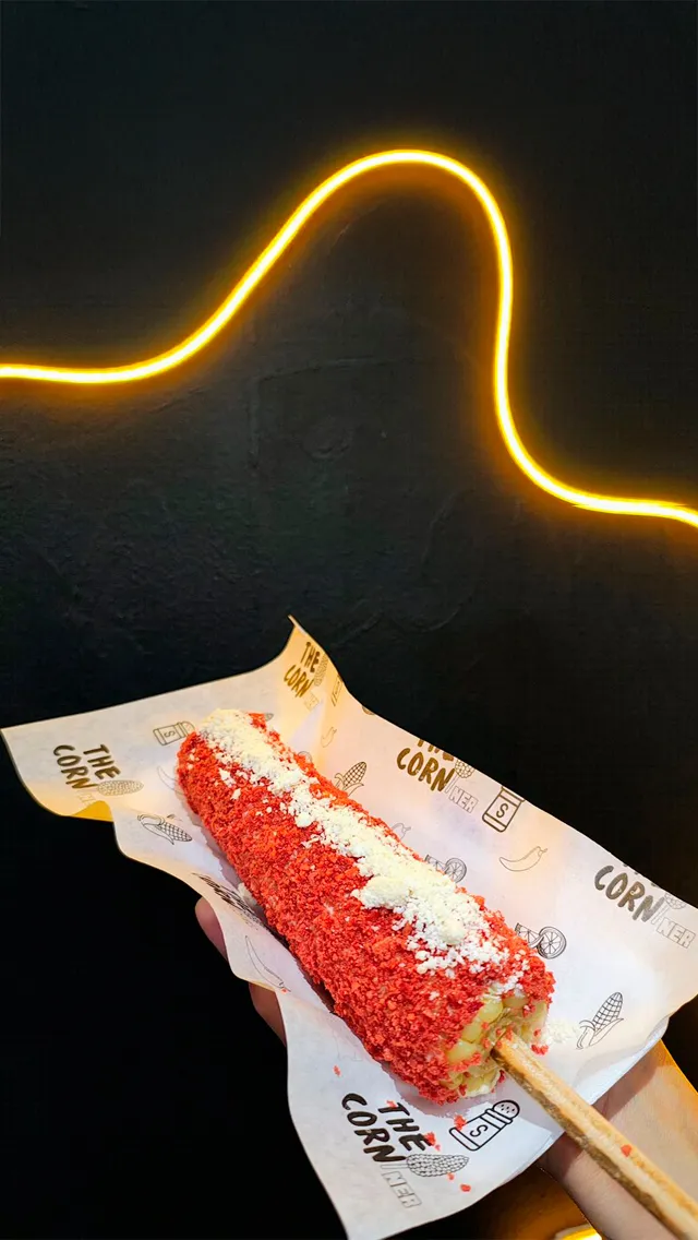 Elote revolcado en papita roja con queso, bajo una luz de neón amarilla