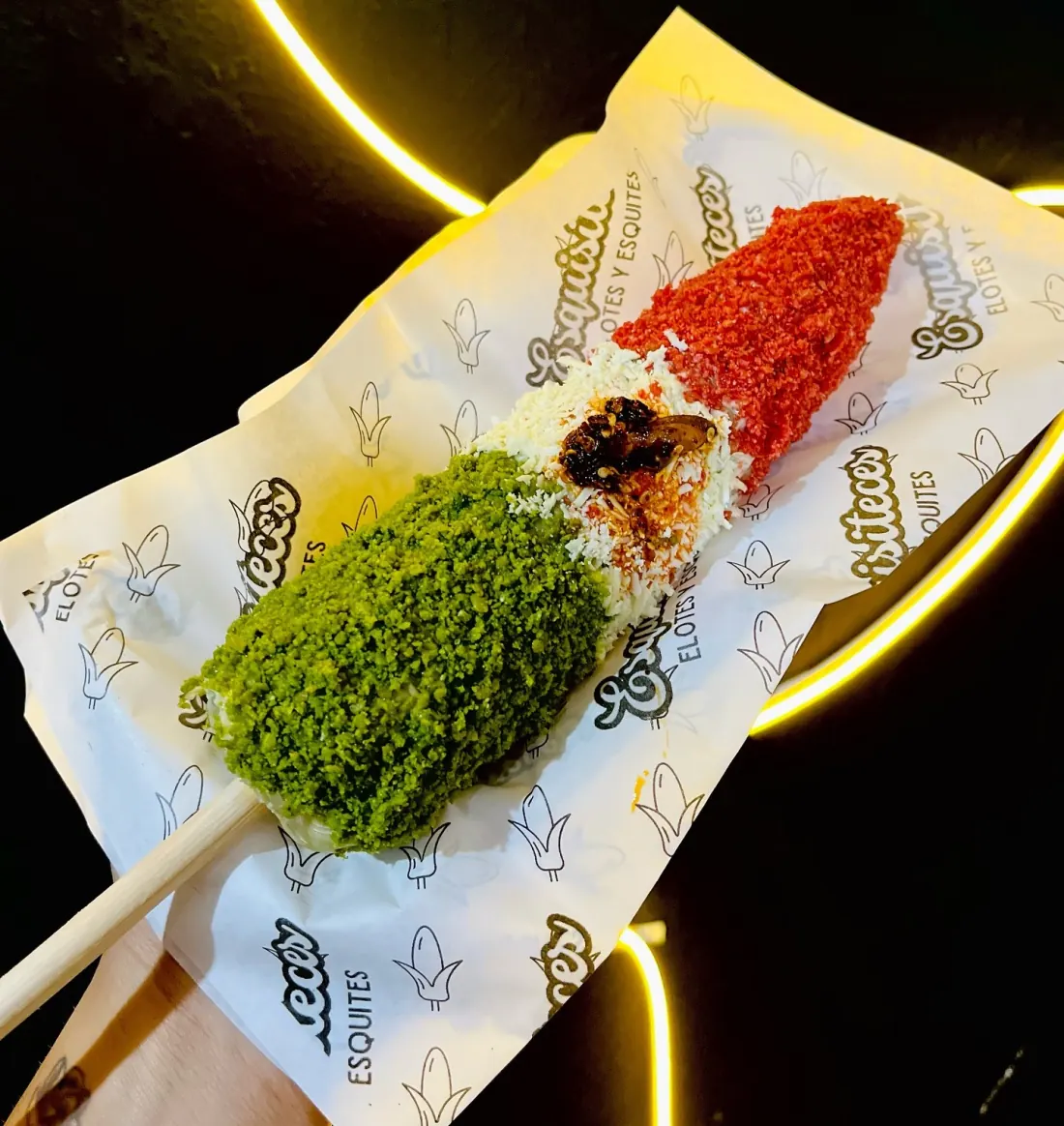 Elote tricolor de Esquisiteces con cobertura verde, queso blanco y cobertura roja, sobre papel con el logo de la marca y luz de neón amarilla