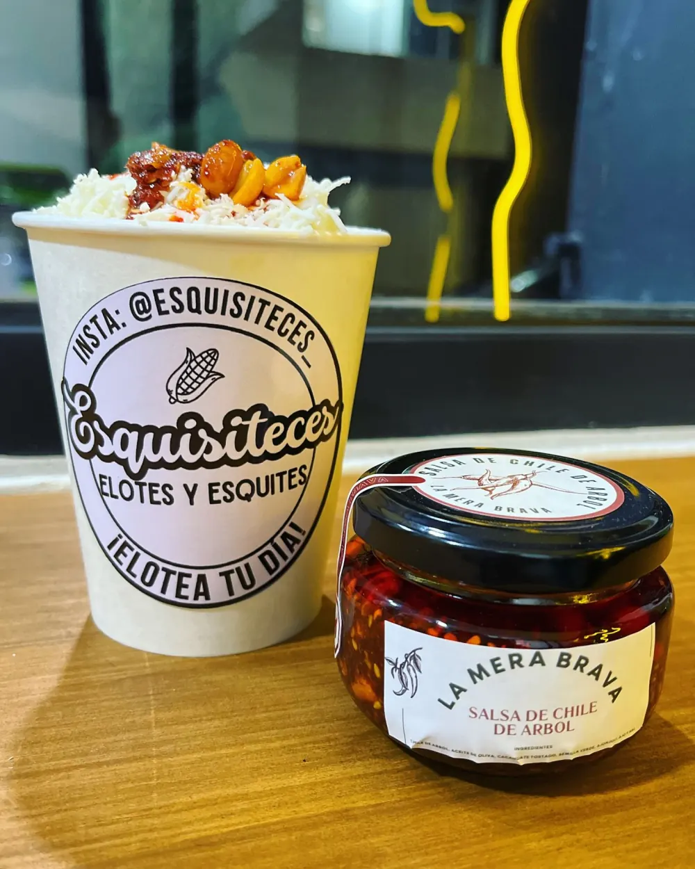 Vaso de esquites de Esquisiteces con queso rallado, cacahuates y salsa de chile, junto a un frasco de salsa de chile de árbol