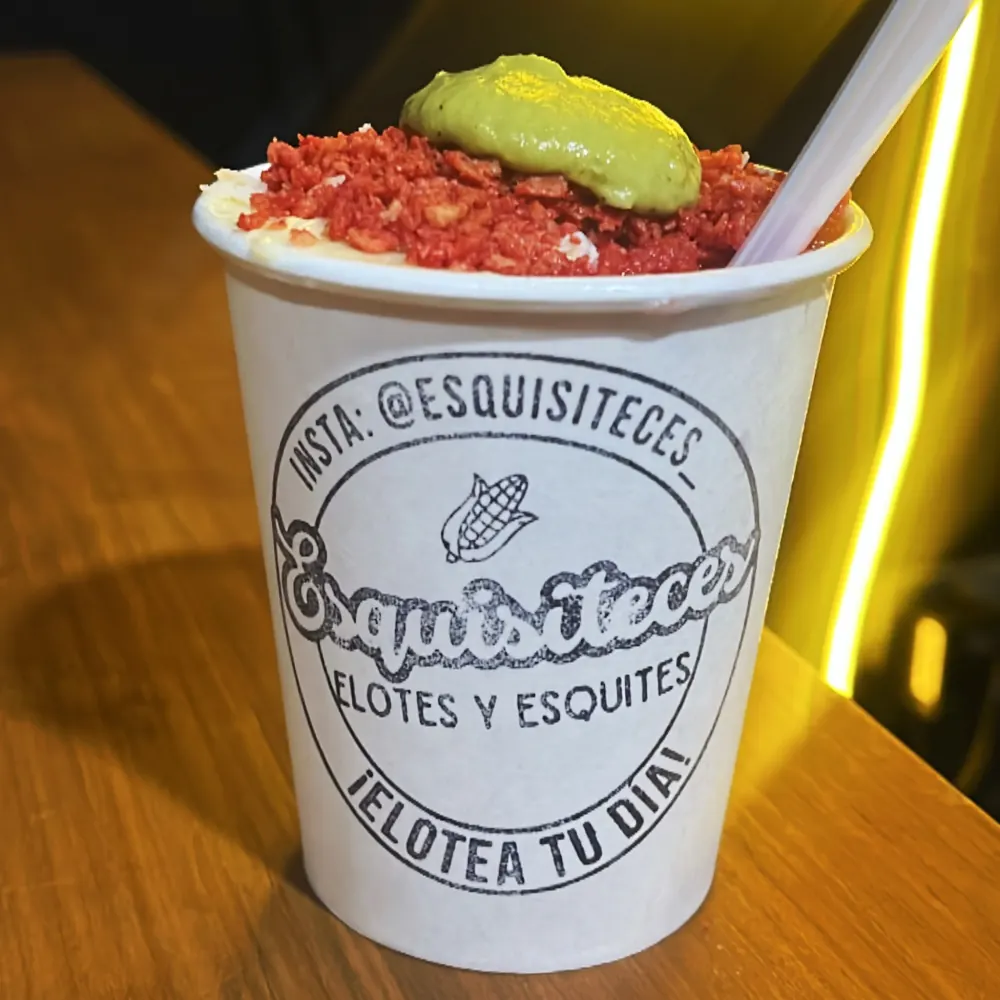 Vaso de esquites de Esquisiteces con cobertura roja crujiente y aderezo verde, con el logo de la marca y un popote